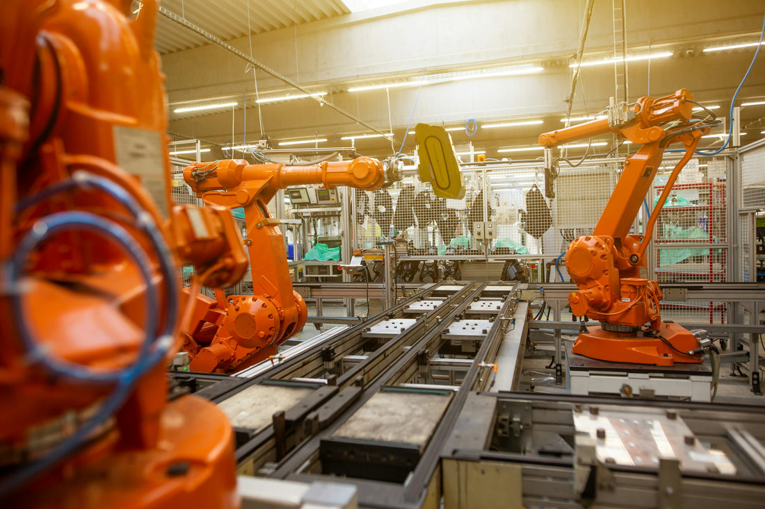 Orange industrial robots on an assembly line, applying masking or coating to a yellow part in a manufacturing plant.