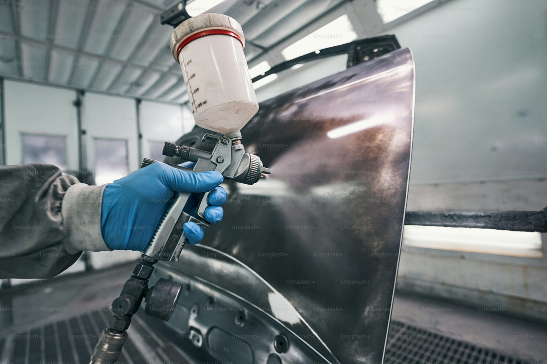 Worker in blue gloves spray coating a metal car part in a booth, illustrating custom masking solutions for industrial finishing.